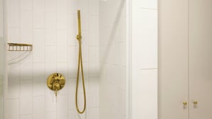 Une douche dotée d’un pommeau et d’un flexible dorés, ainsi que d’un porte-serviettes doré et d’un mur recouvert de carreaux blancs.
