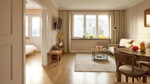 Un salon comprenant un canapé, une table basse et une table à manger. On y trouve également un téléviseur fixé au mur, une fenêtre avec des rideaux et un radiateur.