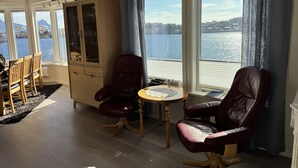 A room with a wooden ceiling, large windows with a view of a lake, a dining area with chairs and a table, and a cabinet with glass doors. - Lofoten Villa by the sea (Vågan)