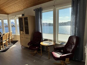 A room with a wooden ceiling, large windows with a view of a lake, a dining area with chairs and a table, and a cabinet with glass doors.