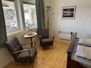 A room with two patterned armchairs, a small round table with a vase and a rose, a floor lamp, a bed with pillows, a radiator, and a framed picture on the wall.