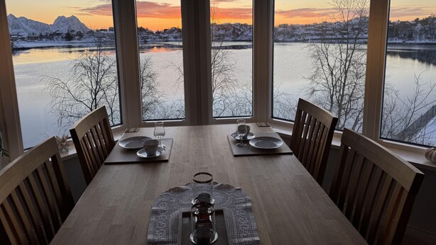 A dining table with chairs, a placemat, and a view of a lake and mountains at sunset. - Lofoten Villa by the sea (Vågan)