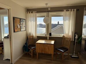 A room with a wooden table and chairs, a corkboard with photos, a window with curtains, and a view of a body of water.