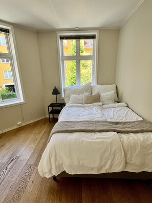 Ein Schlafzimmer mit einem großen Bett, einem Nachttisch, einer Lampe und einem Fenster mit Blick auf Gebäude.