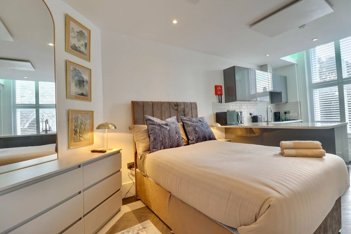 A modern bedroom with a large bed, bedside tables, a desk, and a kitchenette.