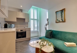 Ett kompakt bostadsutrymme med ett litet kök, en soffa, ett soffbord och ett fönster med persienner.