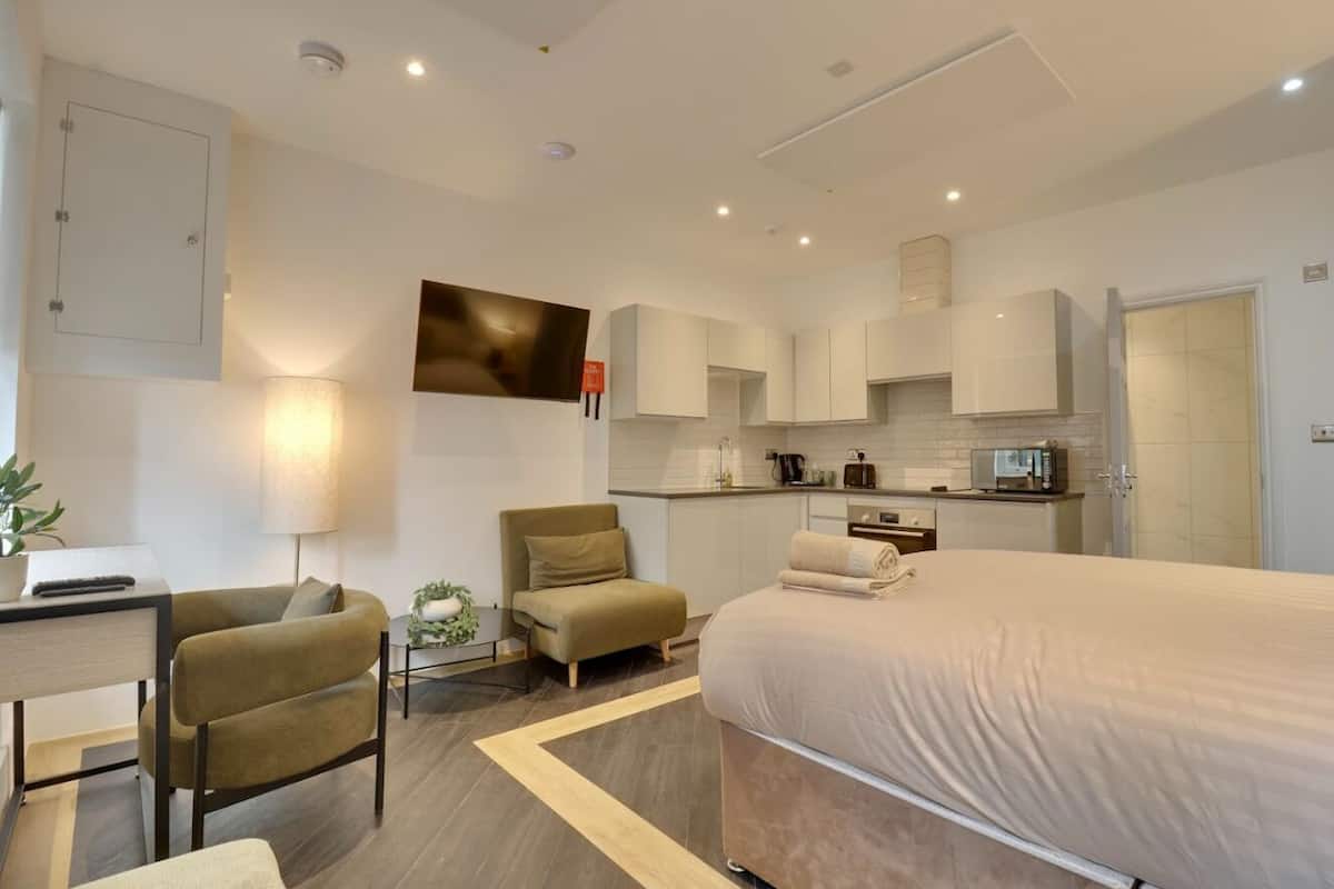 A modern hotel room with a bed, sofa, desk, lamp, and kitchenette.