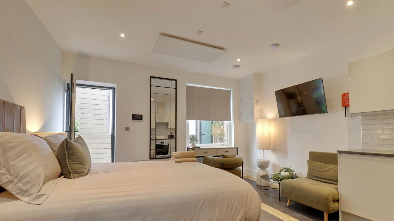 A modern bedroom with a large bed, a TV mounted on the wall, a small table with a lamp, and a window with blinds.
