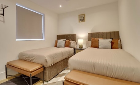 A hotel room with two beds, a bench, a bedside table with a lamp, a window, and a picture on the wall.. QE2 Loft Apartments