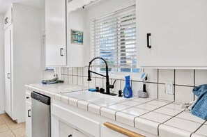 Eine Küche mit weißen Schränken, einem schwarzen Wasserhahn, einer Spülmittelflasche und einem Fenster mit Jalousien.