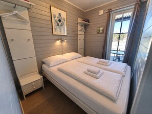 Una cama doble con sábanas blancas, una mesita de noche, un perchero, un cuadro en la pared y una ventana con cortinas.