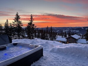 A hot tub in a snowy landscape with a view of a cabin and trees.