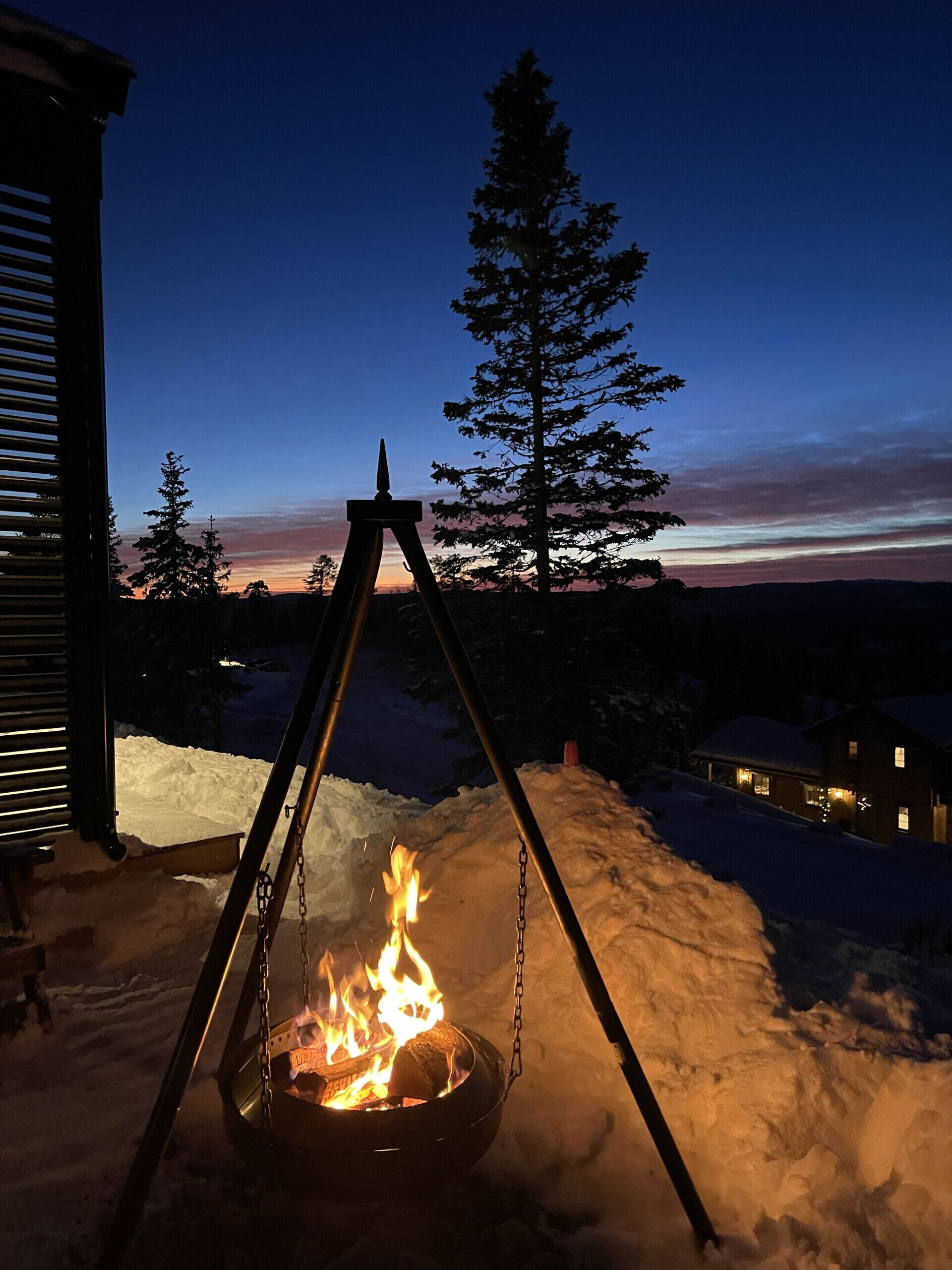 Ein Feuerkorb in einer verschneiten Outdoor-Umgebung, im Hintergrund ein Haus.