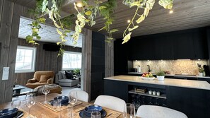 Una cocina moderna con una isla central, un comedor con mesa y sillas de madera, y plantas colgantes.