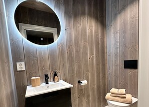 Un baño con pared de madera, un espejo redondo, un lavabo, un inodoro y una toalla sobre el inodoro.