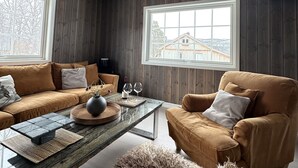 A living room with a sofa, armchair, coffee table, and a view of a wooden house through a window. - Newly renovated house close to everything (Hol)