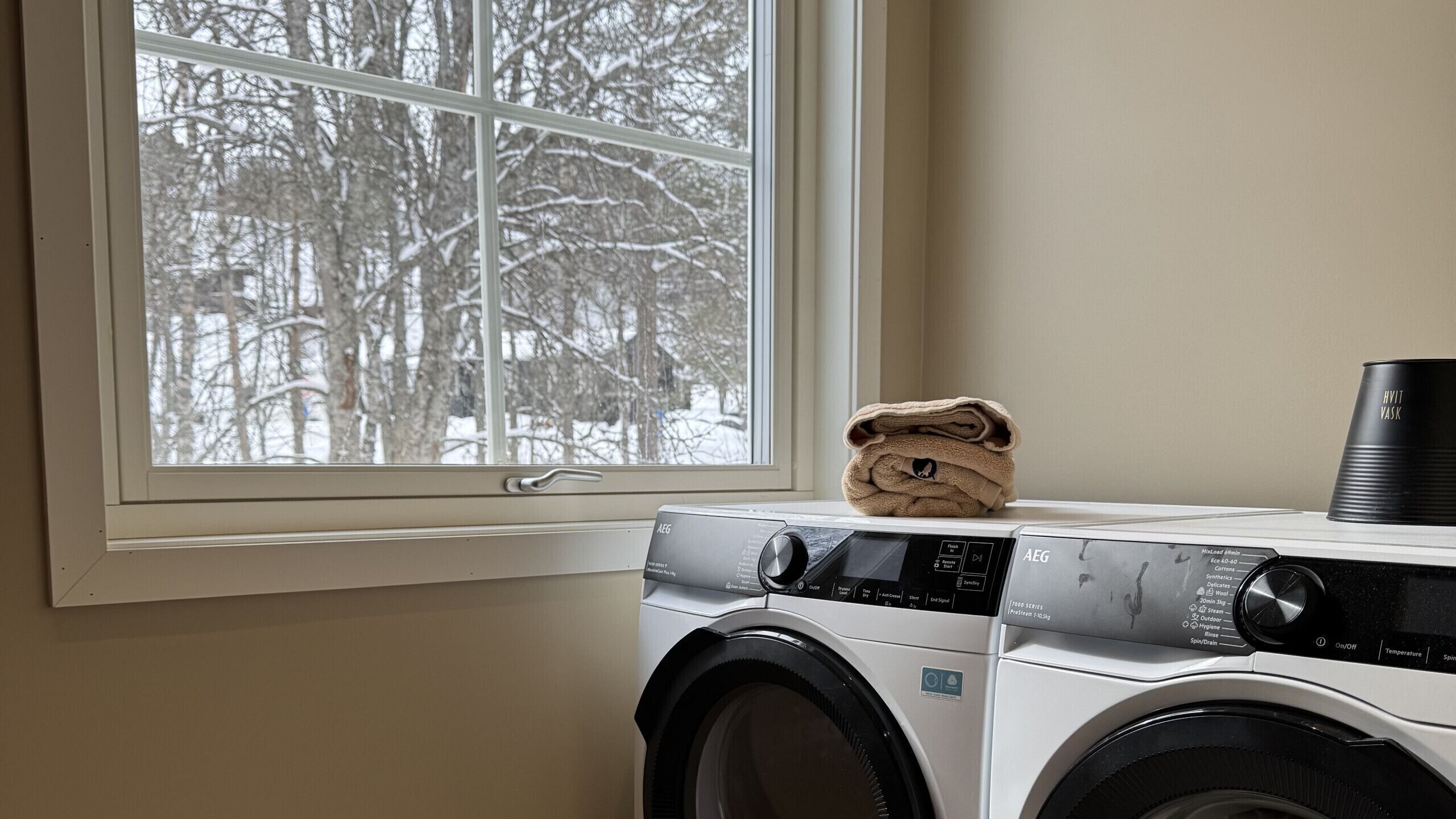Een wasmachine met voorlaadklep, een bedieningspaneel, een raam met uitzicht op besneeuwde bomen en een opgevouwen handdoek erbovenop.