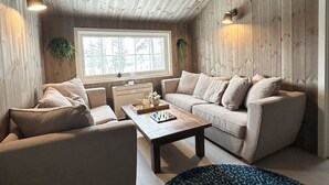 A cozy living room with a wooden ceiling, a large window with a view of trees, a beige sofa, a coffee table with a board game, and a small plant on the wall. - Newly renovated house close to everything (Hol)