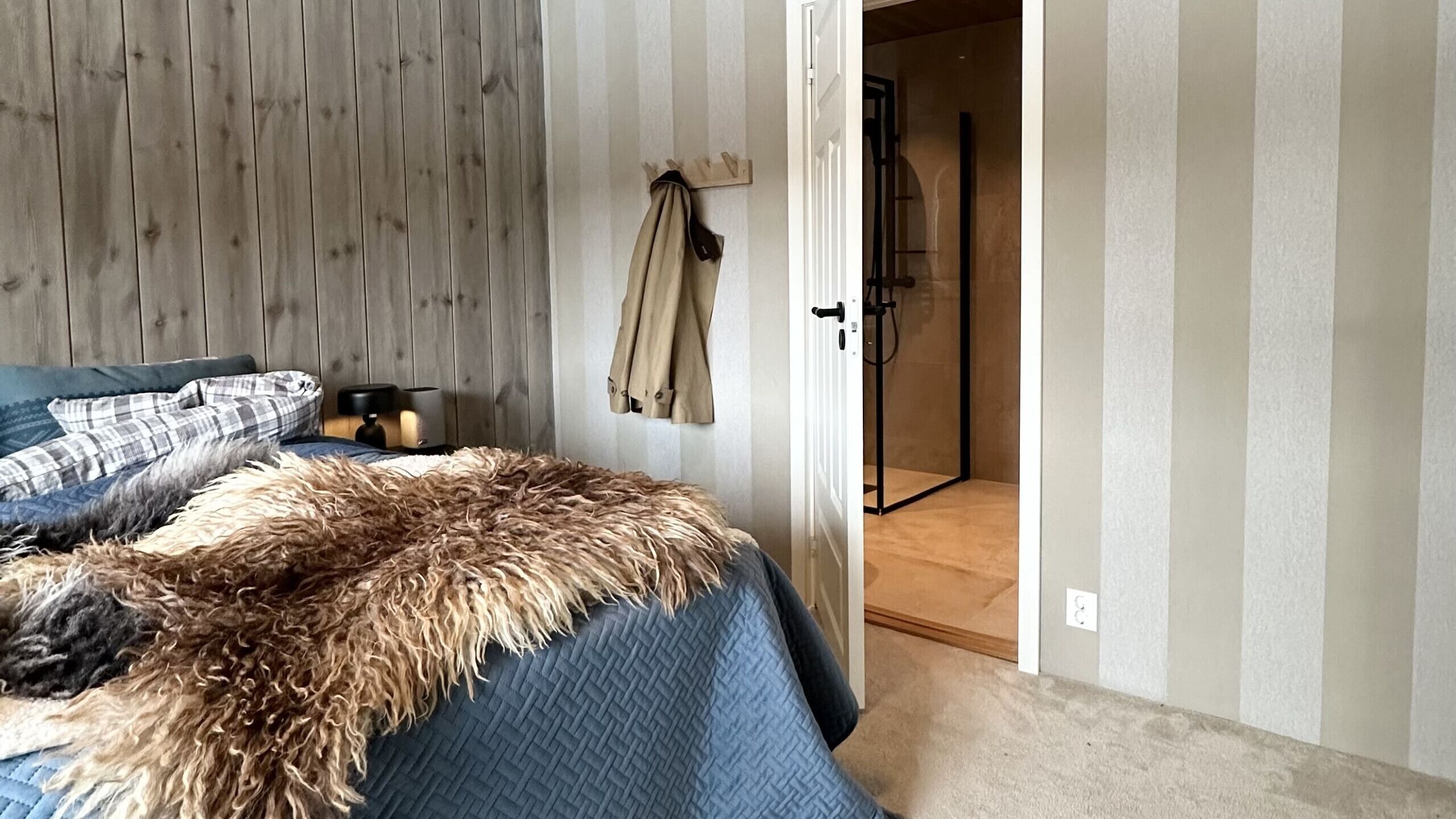 A bedroom with a bed, a wooden headboard, a coat rack, a nightstand with a lamp, and a door leading to a bathroom.