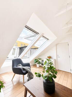 Moderni, avoimen pohjaratkaisun makuuhuone, jossa on kattovalokuvio, tumma nojatuoli, puinen pöytä ja ruukussa oleva kasvi.