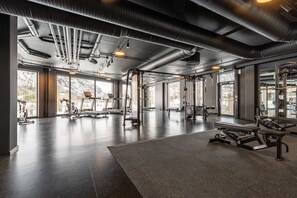 Un gimnasio con equipo de ejercicio, ventanas amplias y un techo alto con conductos a la vista.