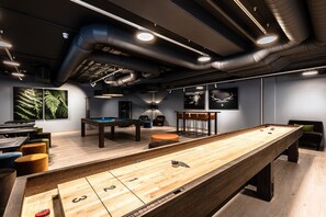 Mesa de billar, mesa de billard y mesa de shuffleboard en una sala moderna con suelo de madera y conductos en el techo.