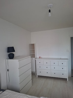 A bedroom with a bed, a white dresser with multiple drawers, a bedside table with a lamp, and a small shelf unit.