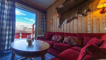 A room with a red sofa, a round wooden table, a mounted animal hide, and a view of mountains through a window.
