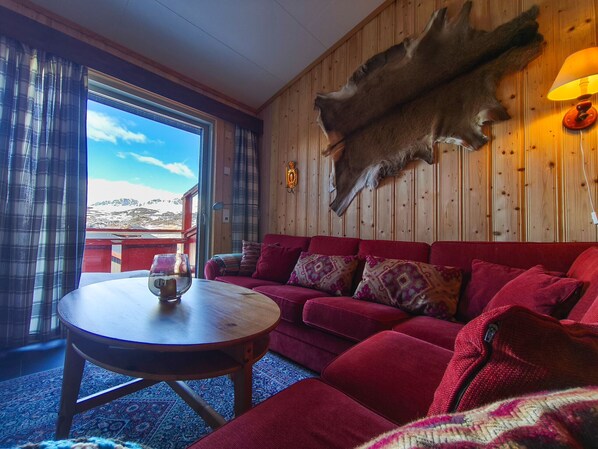 Una habitación con un sofá rojo, una mesa redonda de madera, un trofeo de caza en la pared y vistas a las montañas a través de una ventana.
