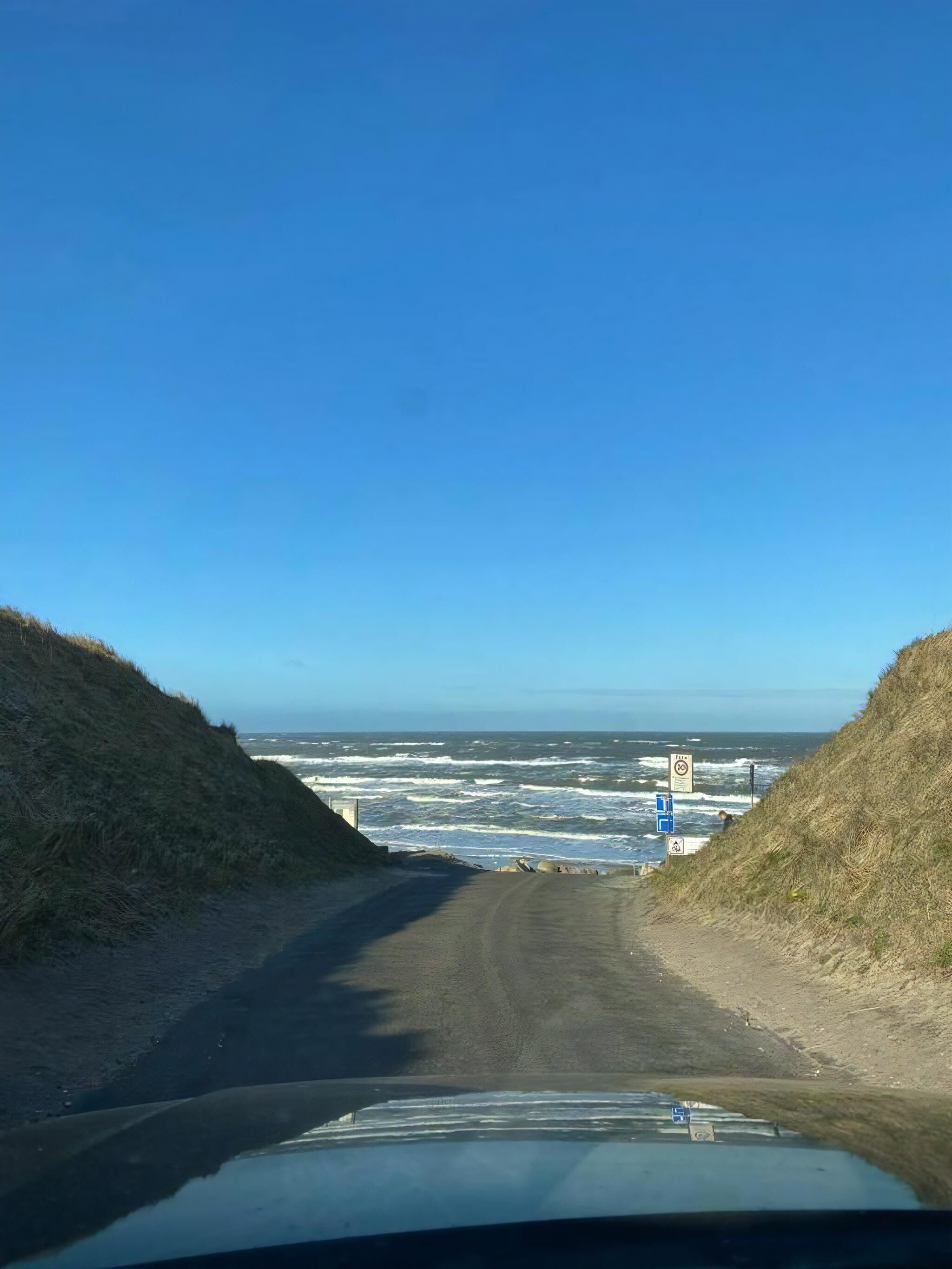 Una strada costiera con vista libera sull'oceano, un segnale che indica divieto di sosta e una targa blu con simboli bianchi.