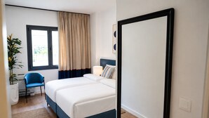 Quarto de hotel com cama, uma cadeira, planta em vaso, janela com cortinas e porta.
