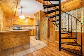 A wooden cabin interior with a kitchen, a central island, a spiral staircase, and a window with a view. - *Spacious 5BD w/ Hot Tub | FirePit + Fireplaces* (Sevierville)