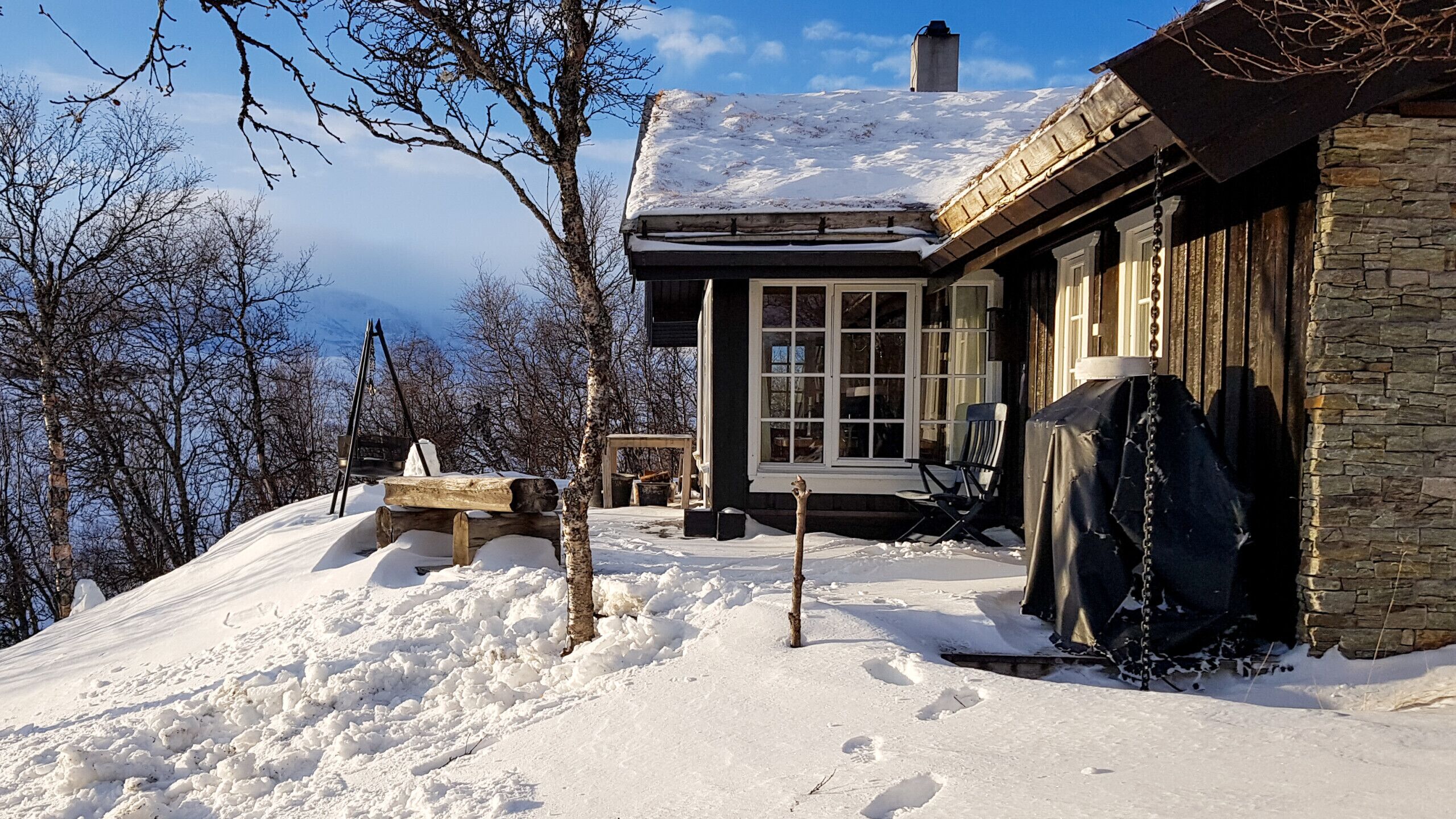 A snow-covered landscape with a stone building featuring a large window and a wooden deck.