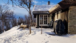 A snow-covered landscape with a stone building featuring a large window and a wooden deck.
