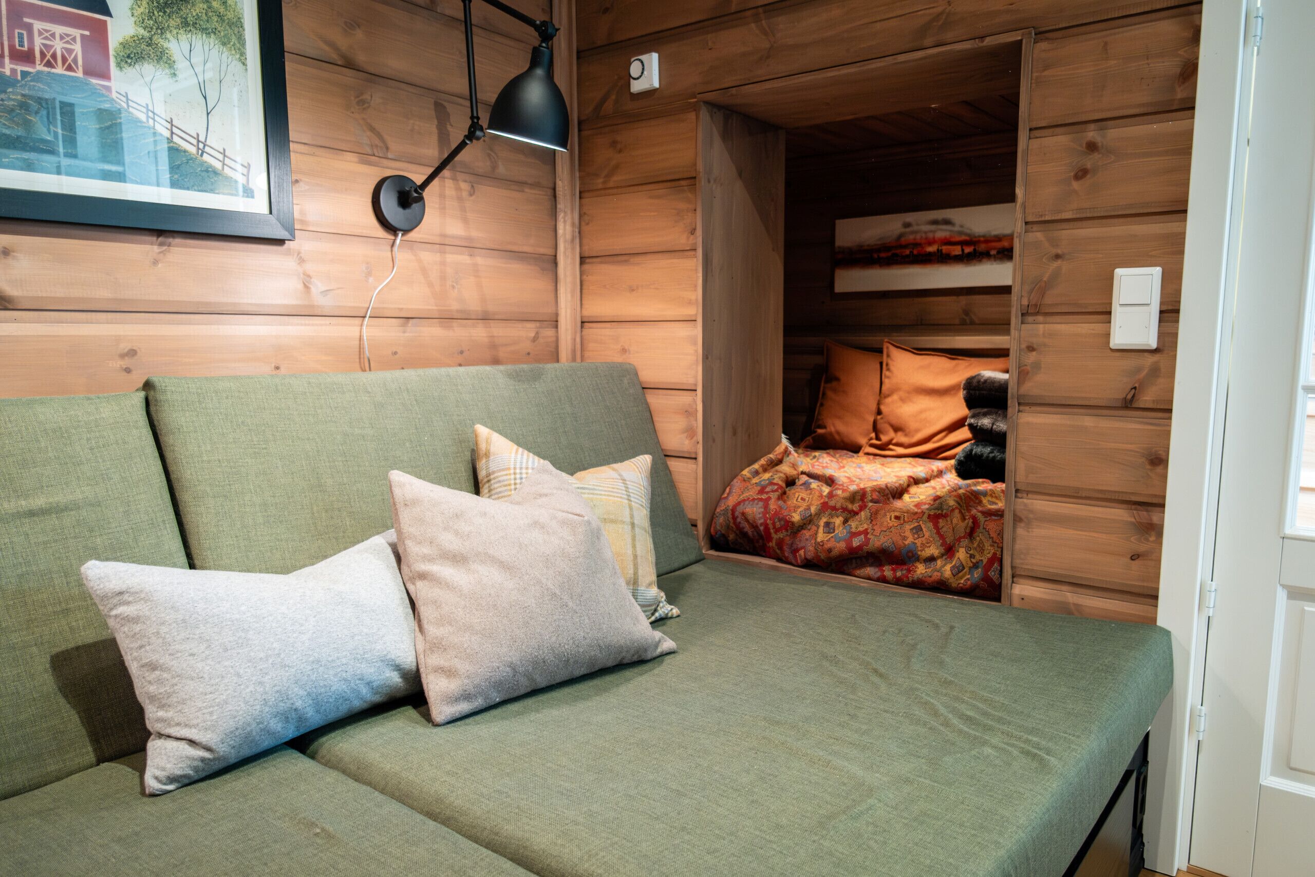 Interior d'una cabana de fusta amb un sofà verd, almoadines, un quadre amb marc i una làmpada.