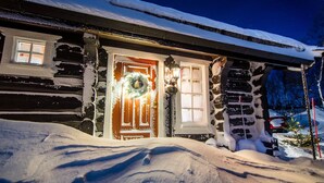 Una cabaña de troncos con la entrada cubierta de nieve, un adorno de corona en la puerta y un muñeco de nieve decorativo.