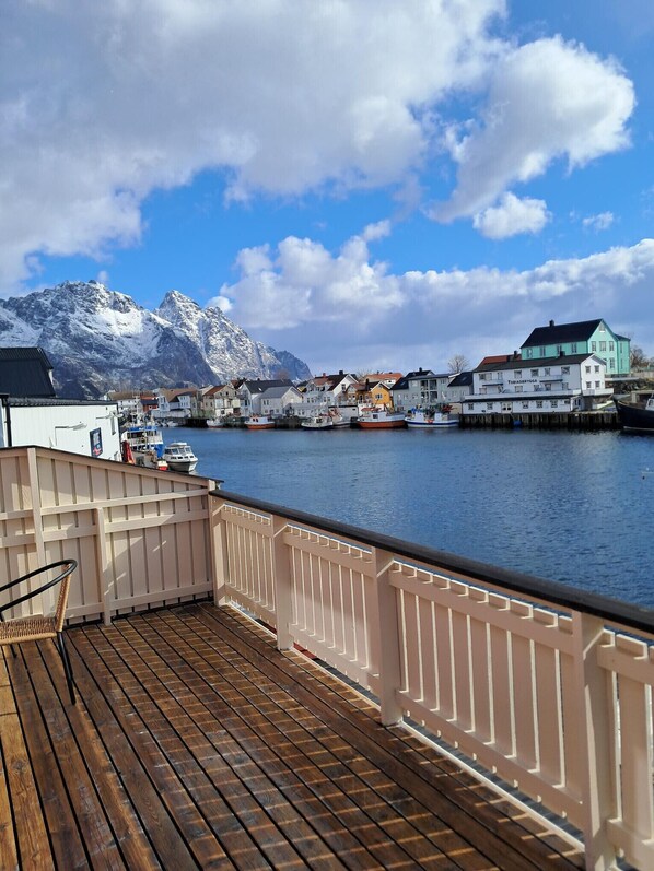 A wooden deck with a railing, a view of a harbor with boats, and buildings with snow-capped mountains in the background.