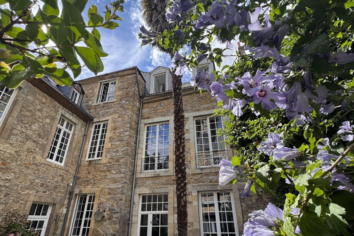 A stone building with multiple windows and a door, surrounded by greenery and flowers.