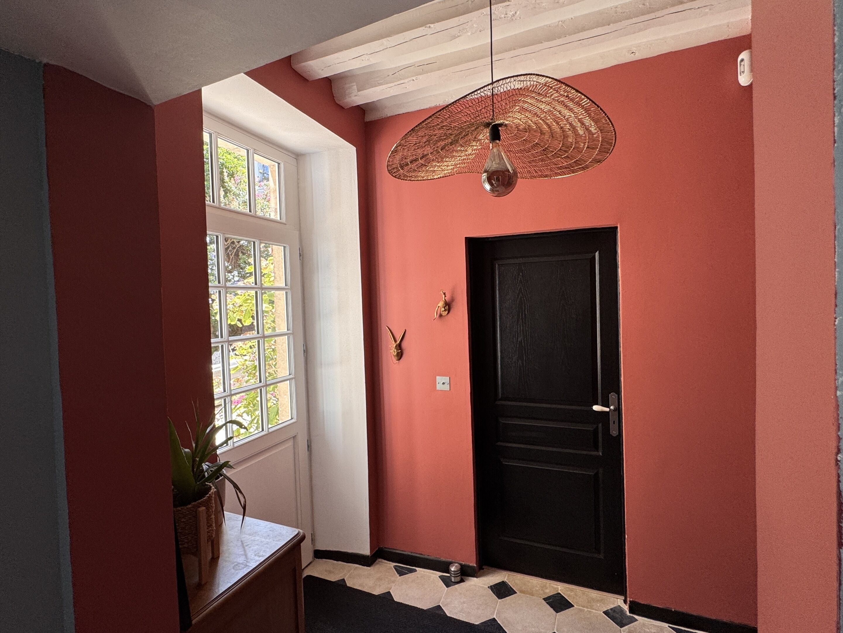 Um corredor com uma porta preta, um lustre pendurado e uma janela com vista para o verde.