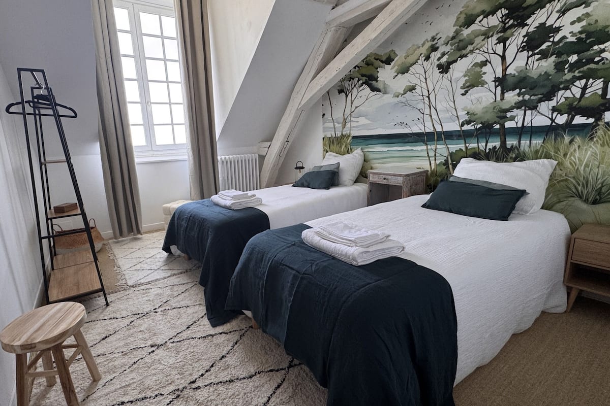 A bedroom with two beds, a radiator, a small table, a chair, and a window with curtains.