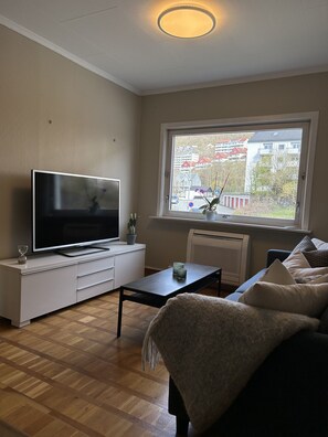 Una sala de estar moderna con un televisor de pantalla plana, una mesa de centro, un sofá y una ventana que ofrece vistas a edificios.