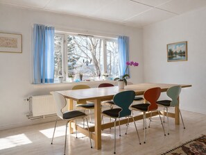 En matplats med ett träbord, fyra stolar, ett fönster med blå gardiner, ett element och en tavla på väggen.