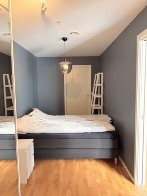 Ein Schlafzimmer mit Bett, Kommode, Spiegel, Deckenleuchte und Tür.