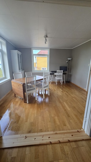 Una habitación amplia con piso de madera, una mesa de comedor con sillas, un ventilador de techo y una ventana que ofrece vistas a un edificio amarillo.