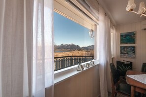 Ein Zimmer mit Fenster und Blick auf eine gebirgige Landschaft, einem Essbereich mit Tisch und Stühlen sowie einer Wandlampe.