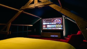 A cozy room with a sloped ceiling, a large screen displaying a TV channel, and red seating.