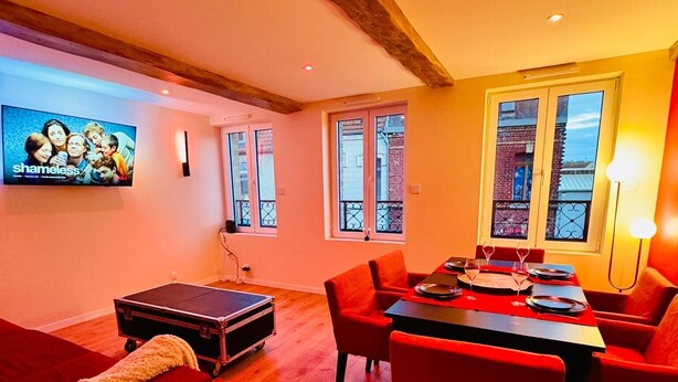 A living room with a red sofa, a dining table with chairs, a television mounted on the wall, and a window with a view of the outside.