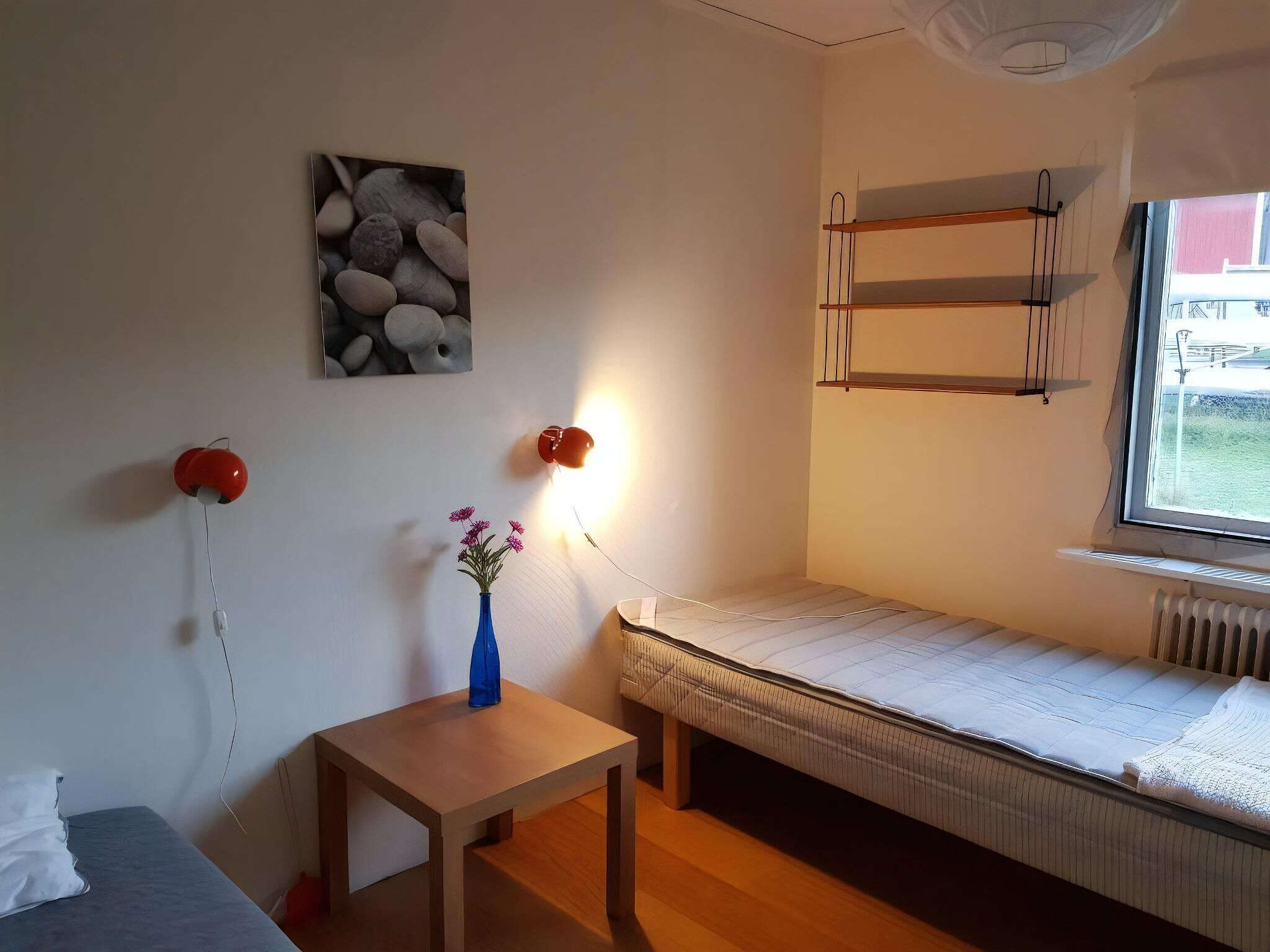 A small, well-lit room with a bed, a bedside table with a vase, a wall-mounted shelf, and a window.