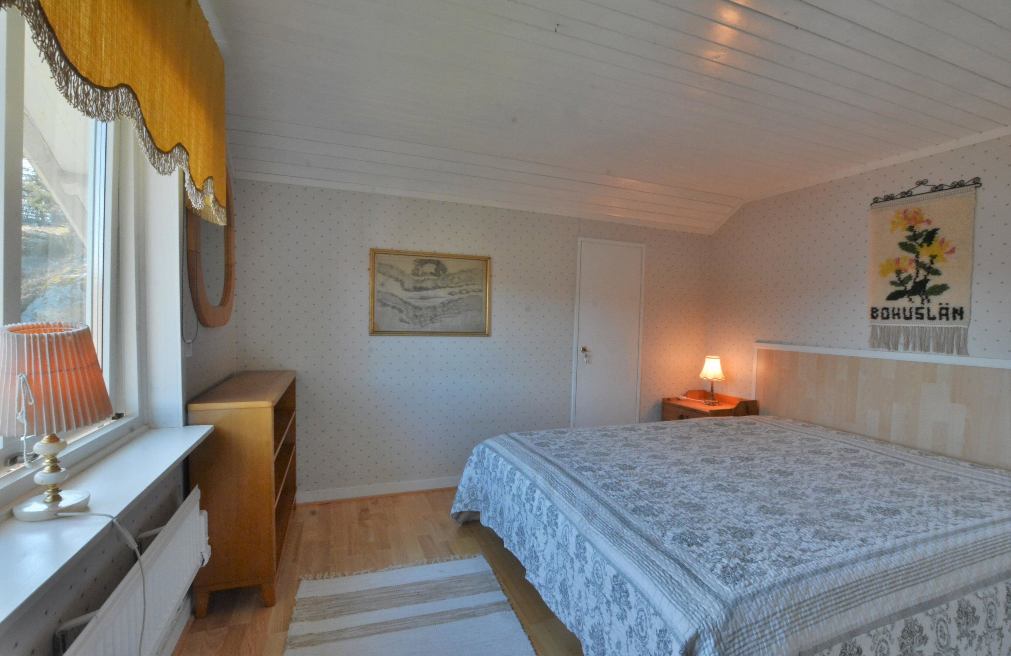 A bedroom with a bed, a nightstand, a dresser, a window with curtains, a wall-mounted lamp, and a framed picture.