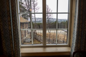 A window with a view of a wooden cabin and a forested area with a mountain in the background.